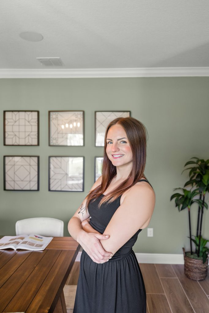 Headshot of Caroline the Realtor, in front of a green background, wood floor, she's angled to the side with her arms crossed and smiling.