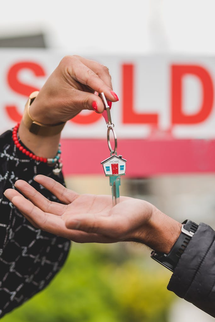 Close-up of hands exchanging house key with colorful SOLD sign in the background.
